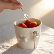 Ceramic mug with sculpted helping hands holding a removable spoon and fork